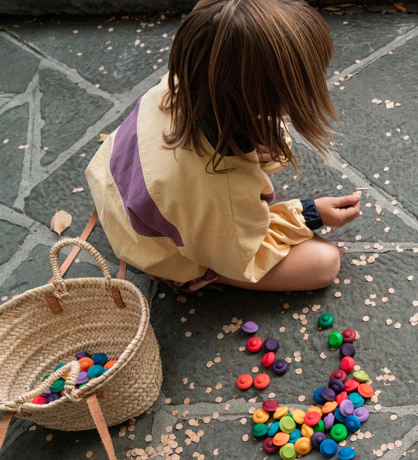 A child playing with the Grapat rainbow flowers mandala pieces. A basket can be seen filled with pieces and some are scattered on the floor around the child. 