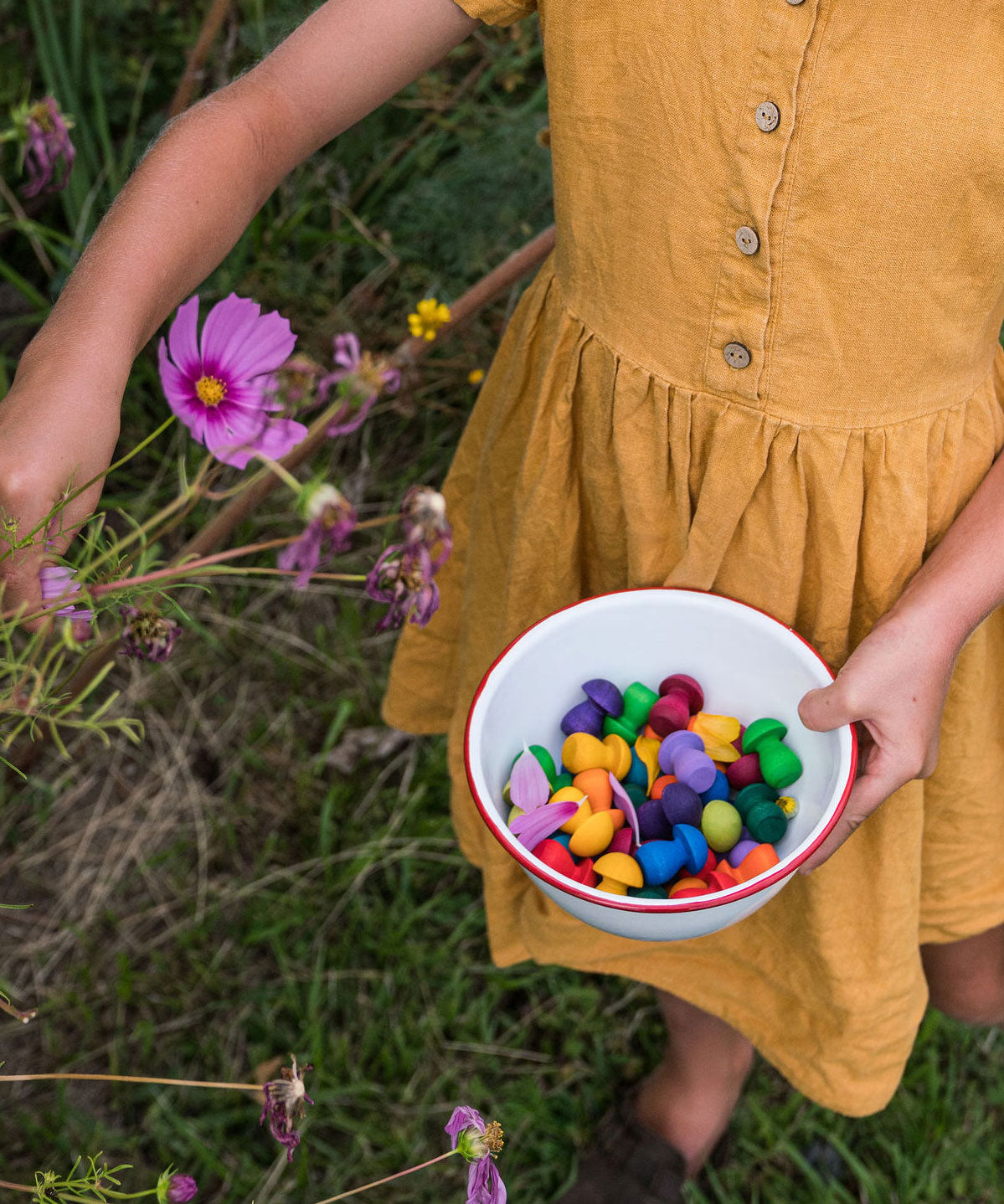 A close up of a chil'ds hand holding a small white enamel bowl filled with the Grapat 36x Mandala Rainbow Mushrooms set. These wooden toys are part of a wide range of open ended toys available at Babipur.  