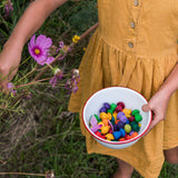 A close up of a chil'ds hand holding a small white enamel bowl filled with the Grapat 36x Mandala Rainbow Mushrooms set. These wooden toys are part of a wide range of open ended toys available at Babipur.  
