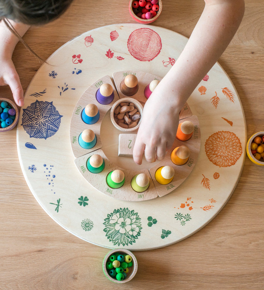 A child placing pieces from teh Grapat perpetual calendar on the Grapat 4 Seasons and Elements Play Platform. A wooden circle shaped platform and stamped with elements and details to represent the four season. These handcrafted items from Grapat are part of a wide range of wooden toys available here at Babipur.