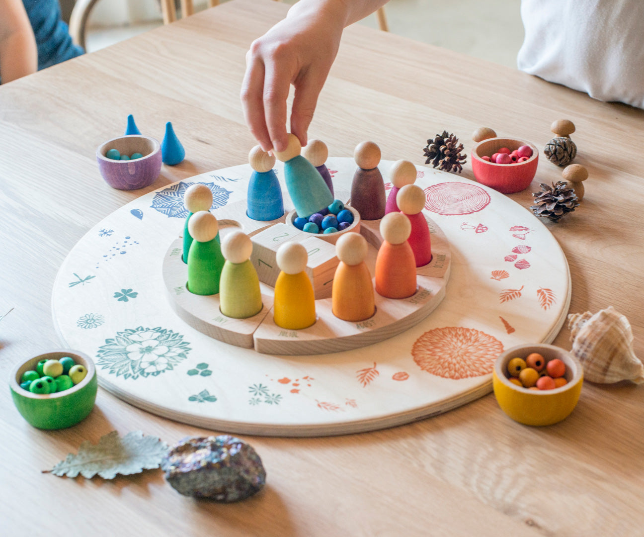 A close up of a child's hand lifting a Grapat Nin peg doll from the Grapat 4 Seasons and Elements Play Platform. A wooden circle shaped platform and stamped with elements and details to represent the four season. These handcrafted items from Grapat are part of a wide range of wooden toys available here at Babipur.