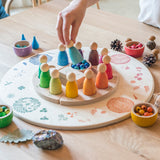A close up of a child's hand lifting a Grapat Nin peg doll from the Grapat 4 Seasons and Elements Play Platform. A wooden circle shaped platform and stamped with elements and details to represent the four season. These handcrafted items from Grapat are part of a wide range of wooden toys available here at Babipur.