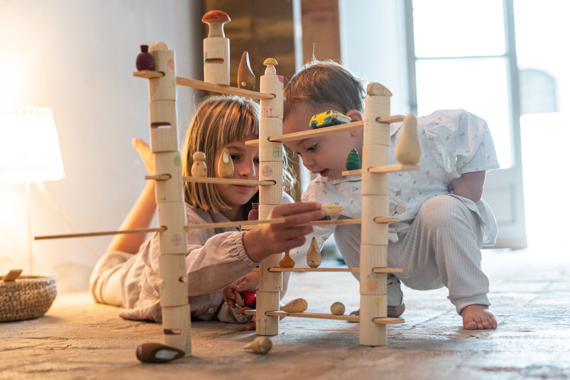 2 children on a stone floor playing with the Grapat woodland set. Showing the tubes and planks stacked into a small world setup with a combination of different Grapat wooden loose parts.
