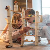 2 children on a stone floor playing with the Grapat woodland set. Showing the tubes and planks stacked into a small world setup with a combination of different Grapat wooden loose parts.