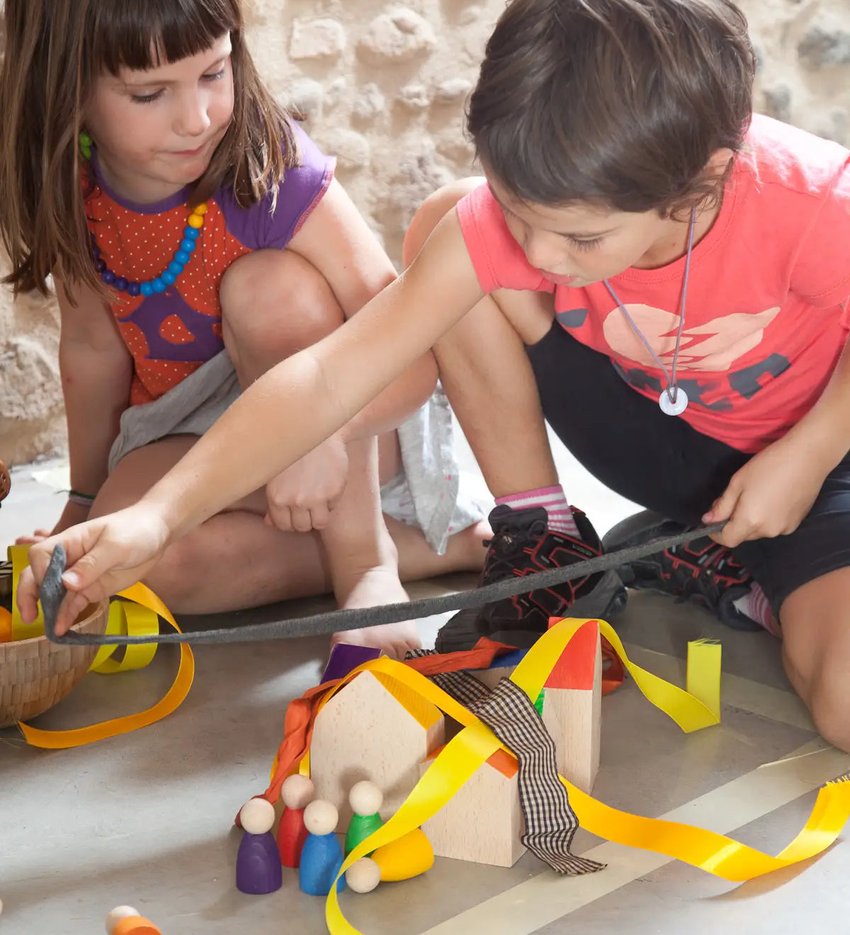 2 children playing with Grapat's wooden house blocks with coloured peg dolls on a concrete surface