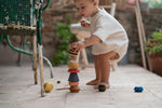 Child outside stacking the pieces from the Grapat serendipity set on a stone floor next to a garden chair. Features a handcrafted brown, light pink, blue yellow orange and striped pieces. 