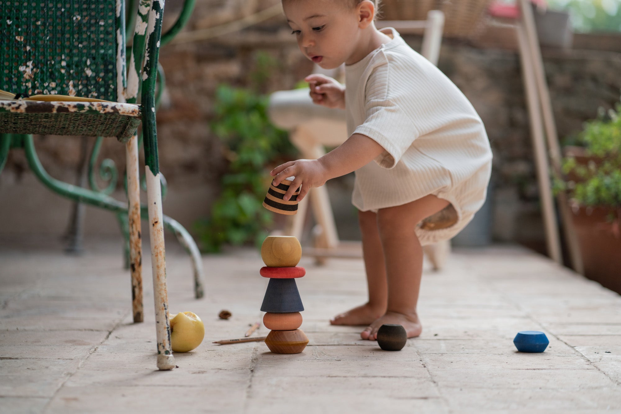 Child outside stacking the pieces from the Grapat serendipity set on a stone floor next to a garden chair. Features a handcrafted brown, light pink, blue yellow orange and striped pieces. 