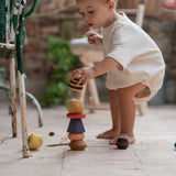 Child outside stacking the pieces from the Grapat serendipity set on a stone floor next to a garden chair. Features a handcrafted brown, light pink, blue yellow orange and striped pieces. 