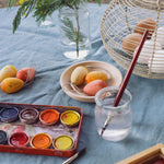 The three eggs from the Grapat Handmade Sunrise Eggs Wooden Easter Decorations set  in a small round wooden dish. A tin containing a watercolour paint pallet can be seen in front of them with a small pit of water and a paintbrush. 