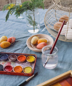 The three eggs from the Grapat Handmade Sunrise Eggs Wooden Easter Decorations set  in a small round wooden dish. A tin containing a watercolour paint pallet can be seen in front of them with a small pit of water and a paintbrush. 