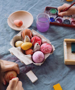 The Grapat Handmade Sunrise Eggs Wooden Easter Decorations shown placed in a cardboard egg tray next to 3 real eggs that have been painted in various colours. 