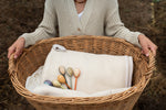 Adult holding a large woven basket with a cream blanket inside with the 6 different Grapat twin soul handcrafted and painted wooden puppets on top of the blanket. 