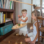 2 children sitting on a terracotta floor playing with the Grapat wooden pendulum set resting on a wooden chair and bookshelf with wooden blocks surrounding them and on the pendulum.