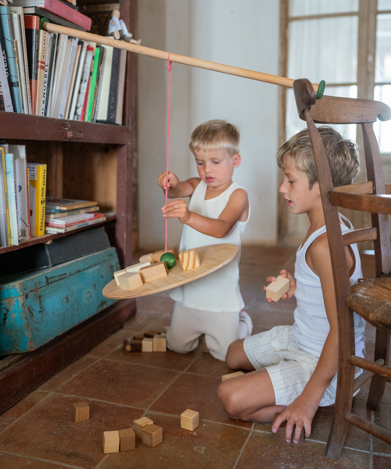 2 children sitting on a terracotta floor playing with the Grapat wooden pendulum set resting on a wooden chair and bookshelf with wooden blocks surrounding them and on the pendulum.