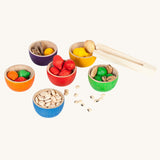 The Grapat Wooden Toy Bowls and  Acorns Set filled with the acorn shapes and dried pulses and small slices of wood. A set of 6 coloured bowls with wooden acorn shapes in matching pieces. These wooden toys are part of a wide range of open ended toys available at Babipur. 