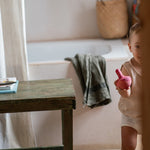 Child holding the Grapat wooden pink chill wobbly bird next to a wooden table in the bathroom on a stone floor