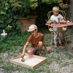 A child gardening on a garden table and another child playing with the Grapat wooden yellow flowing bird in the Grapat wooden freeplay tray filled with sand on the gravel floor.