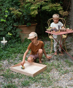 A child gardening on a garden table and another child playing with the Grapat wooden yellow flowing bird in the Grapat wooden freeplay tray filled with sand on the gravel floor.