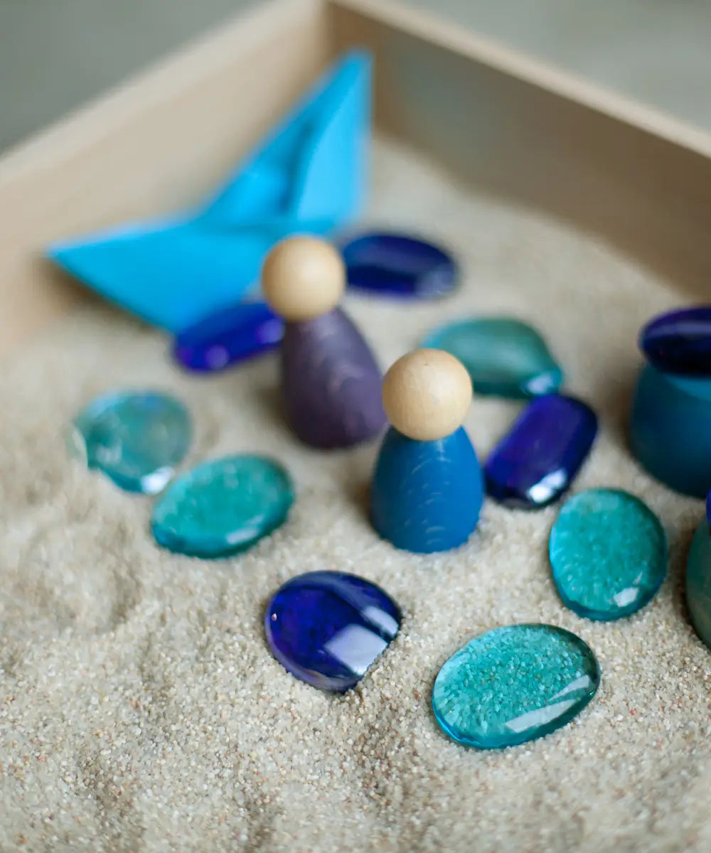 Blue wooden peg dolls and other loose pieces in sand inside a Grapat freeplay wooden tray