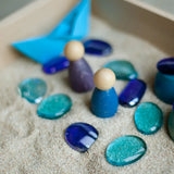 Blue wooden peg dolls and other loose pieces in sand inside a Grapat freeplay wooden tray