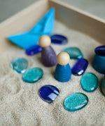 Blue wooden peg dolls and other loose pieces in sand inside a Grapat freeplay wooden tray