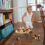 Child standing next to the Grapat wooden pendulum set balancing on a wooden chair and bookshelf with wooden blocks on the wooden balance board above a terracotta floor