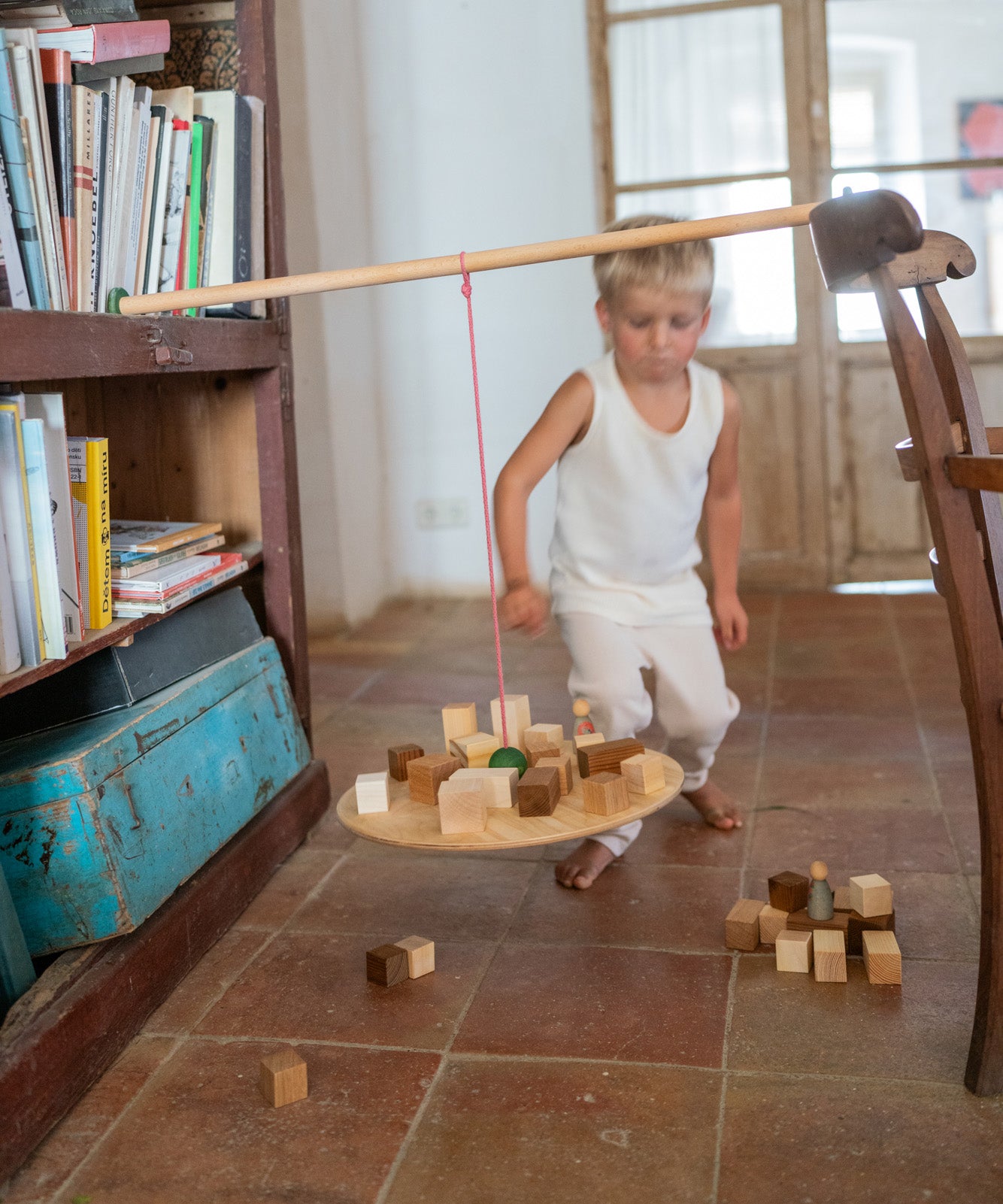 Child standing next to the Grapat wooden pendulum set balancing on a wooden chair and bookshelf with wooden blocks on the wooden balance board above a terracotta floor