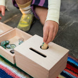 A close up of a child posting a Nin into the Grapat Permanence Box. The wooden box that has two slots in the top one for coins and one circular that will fit small toys such as Nin peg dolls. This wooden box is part of a wide range of open ended toys available at Babipur. 