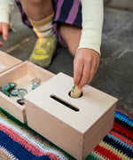A close up of a child posting a Nin into the Grapat Permanence Box. The wooden box that has two slots in the top one for coins and one circular that will fit small toys such as Nin peg dolls. This wooden box is part of a wide range of open ended toys available at Babipur. 
