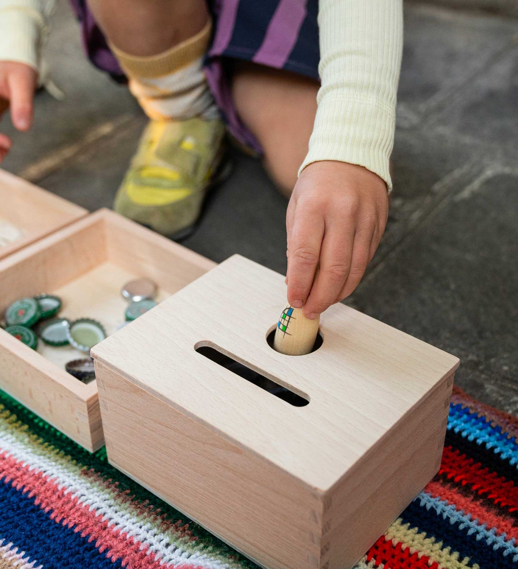 A close up of a child posting a Nin into the Grapat Permanence Box. The wooden box that has two slots in the top one for coins and one circular that will fit small toys such as Nin peg dolls. This wooden box is part of a wide range of open ended toys available at Babipur. 