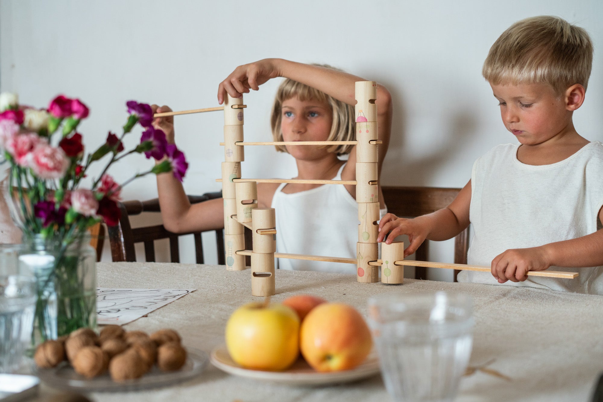 2 children sitting next to a table playing with the handcrafted, painted tubes and planks from the woodland set stacked on a table next to apples and a vase of flowers. 