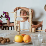 2 children sitting next to a table playing with the handcrafted, painted tubes and planks from the woodland set stacked on a table next to apples and a vase of flowers. 