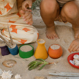 Children playing with the Grapat Wooden Rainbow Sorting Pots on teh sand. Wooden pots in a mix of shapes in different colours.