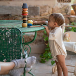 Child standing on a stone table next to a garden table playing with the Grapat wooden serendipity set showing the different shaped pieces stacked on the table. Features a brown, light pink, blue yellow orange and striped pieces. 
