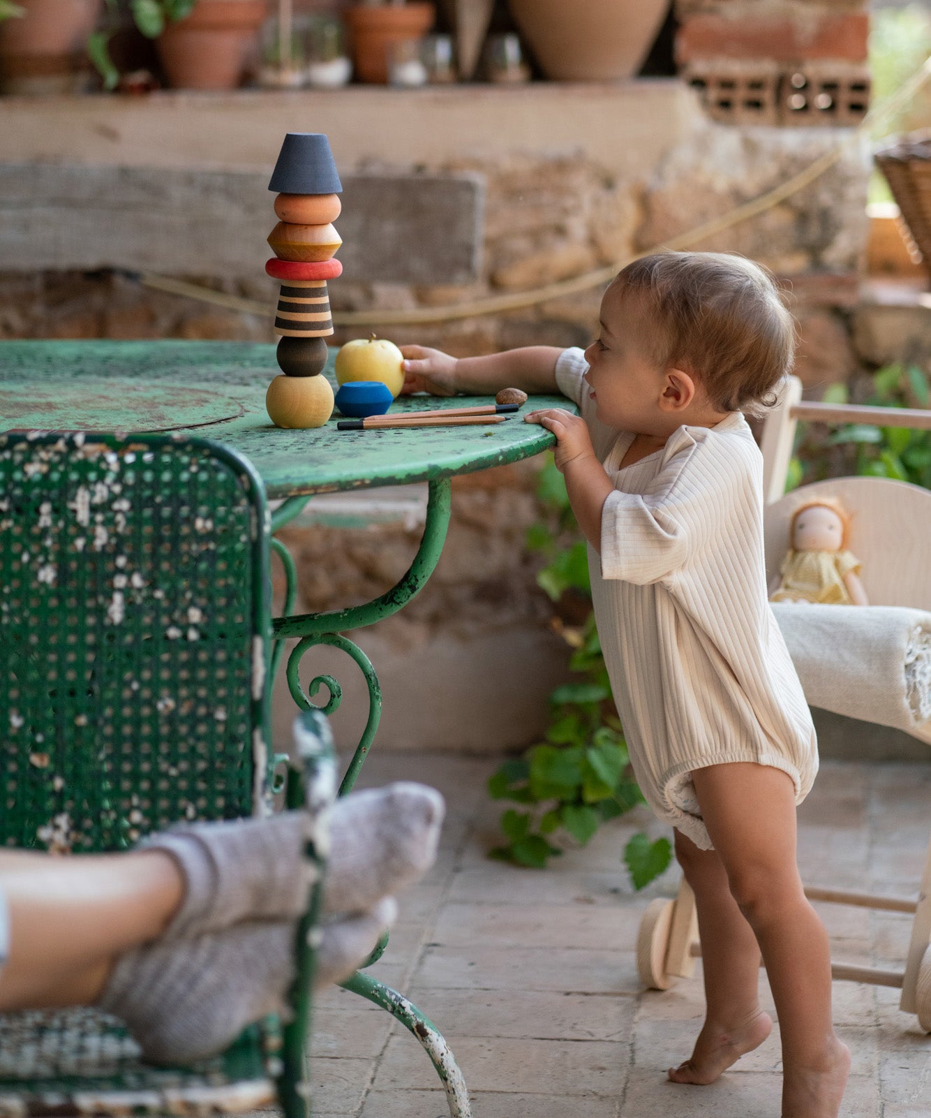 Child standing on a stone table next to a garden table playing with the Grapat wooden serendipity set showing the different shaped pieces stacked on the table. Features a brown, light pink, blue yellow orange and striped pieces. 