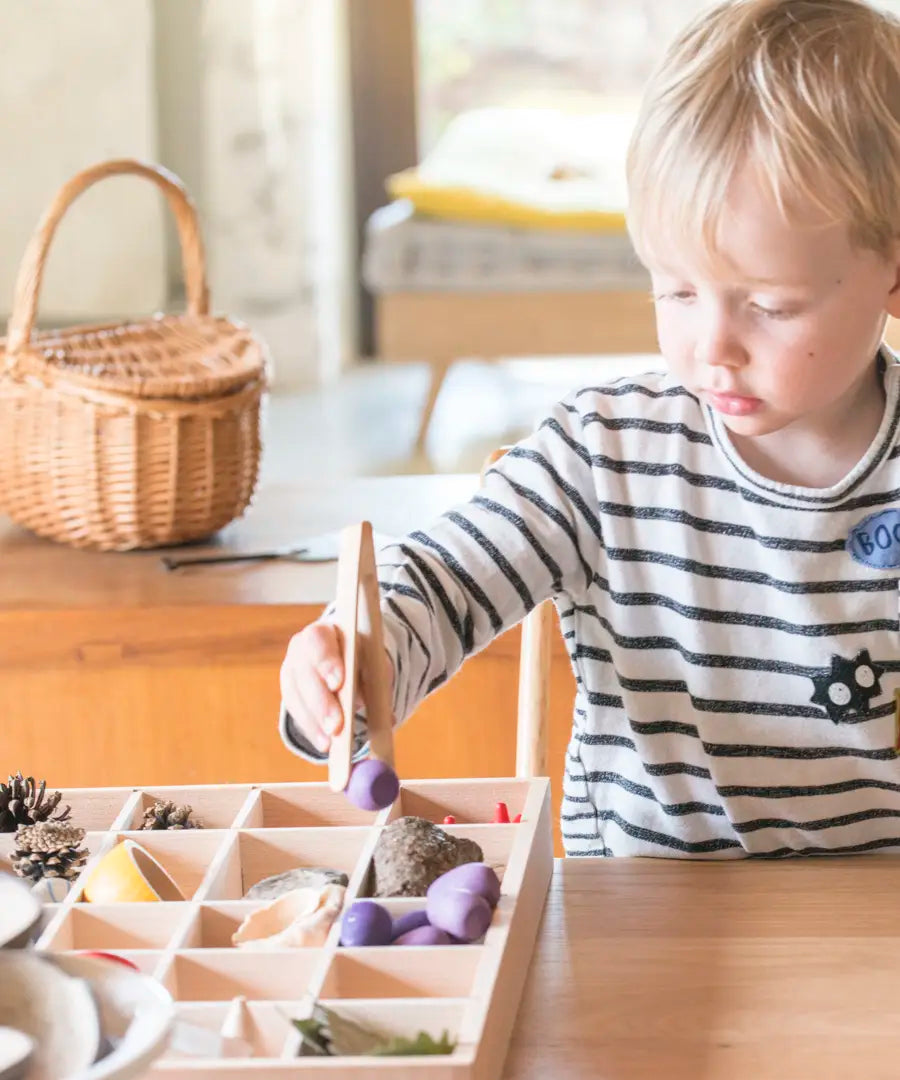 Child standing next to a a Grapat tinker tray and using tongs to fill a compartment with loose wooden pieces