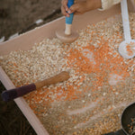 A child looking down at one of the Grapat Wooden Sensory Play Tools. The tool is being held by an adult who is different sized food items poked into the holes of the measuring tool