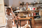 Child standing next to a wooden stool holding a orange and light grey Grapat handcrafted painted wooden puppet with 4 wooden puppets on a wooden stand on the kitchen table and a child sitting on a wooden chair.