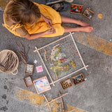 A child sitting on the ground with a Grapat wooden Weaving Frame next to them The weaving frame has been decorated with bits of greenery, wool and flowers.