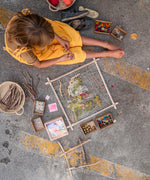 A child sitting on the ground with a Grapat wooden Weaving Frame next to them The weaving frame has been decorated with bits of greenery, wool and flowers.