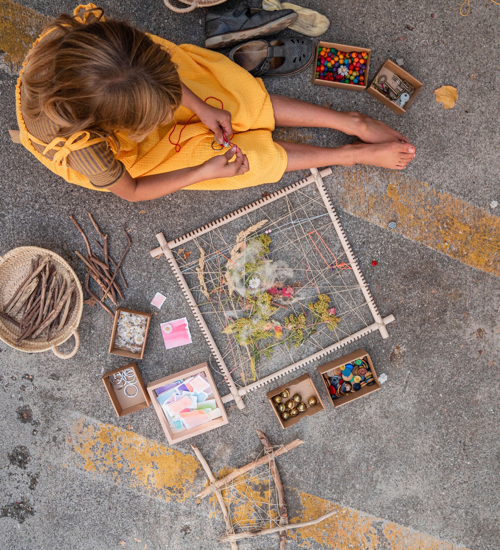 A child sitting on the ground with a Grapat wooden Weaving Frame next to them The weaving frame has been decorated with bits of greenery, wool and flowers.