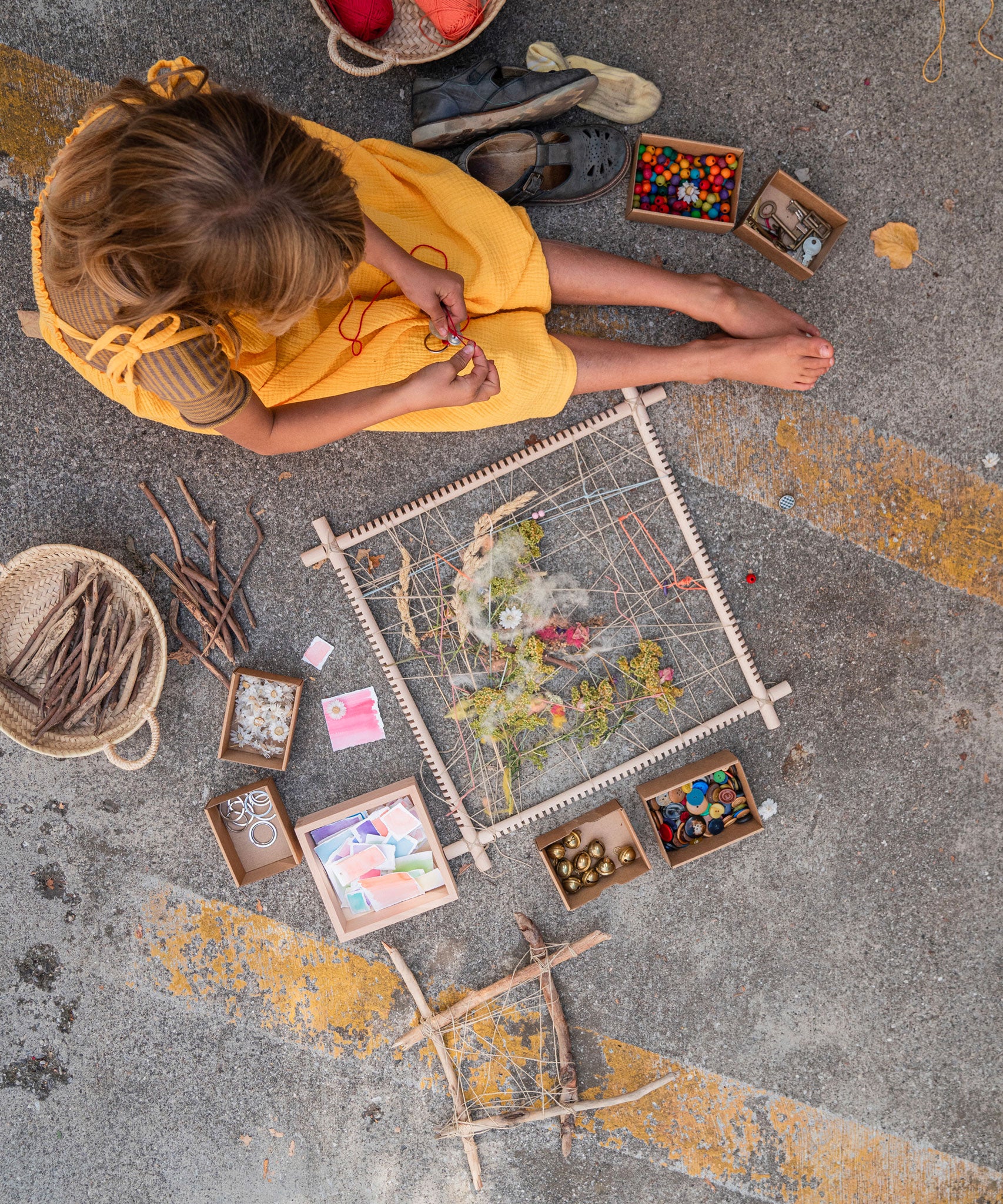 A child sitting on the ground with a Grapat wooden Weaving Frame next to them The weaving frame has been decorated with bits of greenery, wool and flowers.