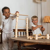 2 children sitting next to a table playing with the handcrafted, painted tubes and planks from the woodland set stacked on a wooden table