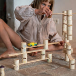 Child sitting on a stone floor stacking the wooden tubes from the Grapat woodland set and balancing a wooden plank on top of another plank. 