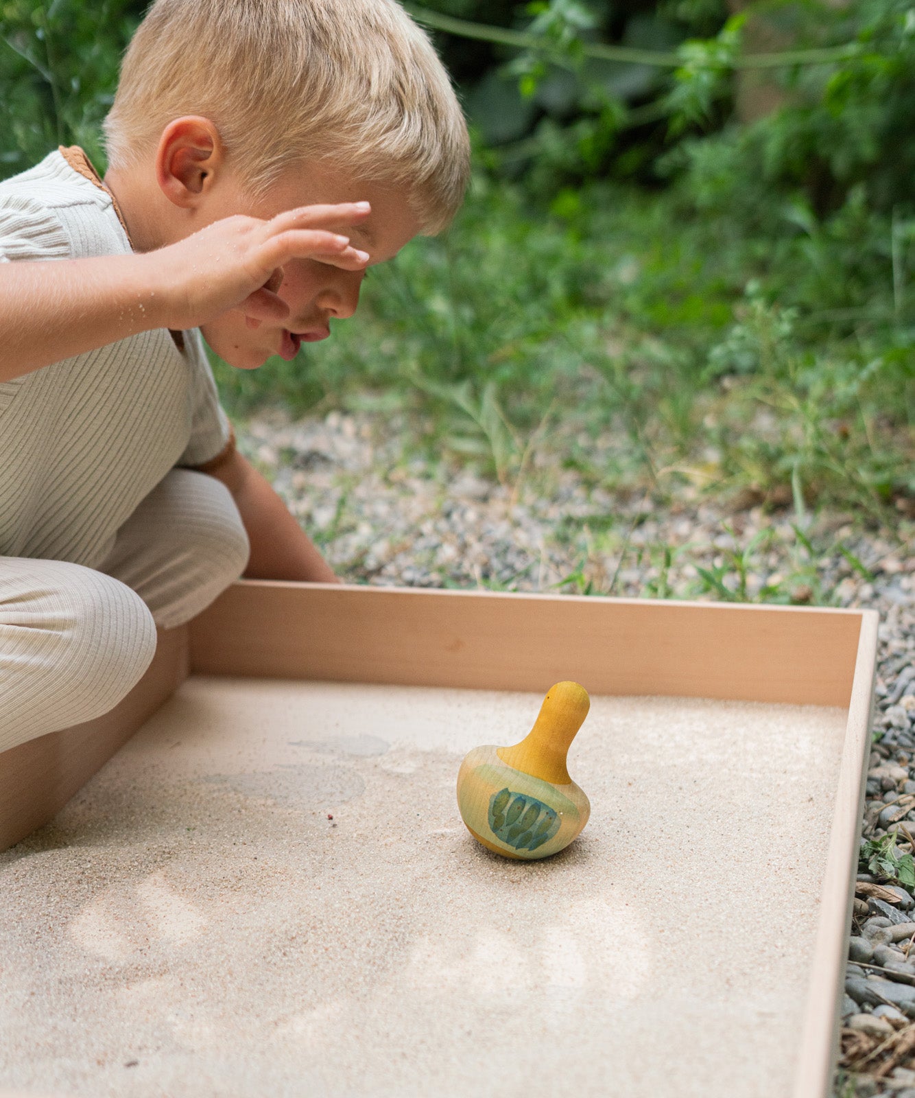 A child in the garden playing with the Grapat wooden yellow flowing bird in a Grapat freeplay tray filled with sand on a gravel floor. Showing the blue and green feather detailing