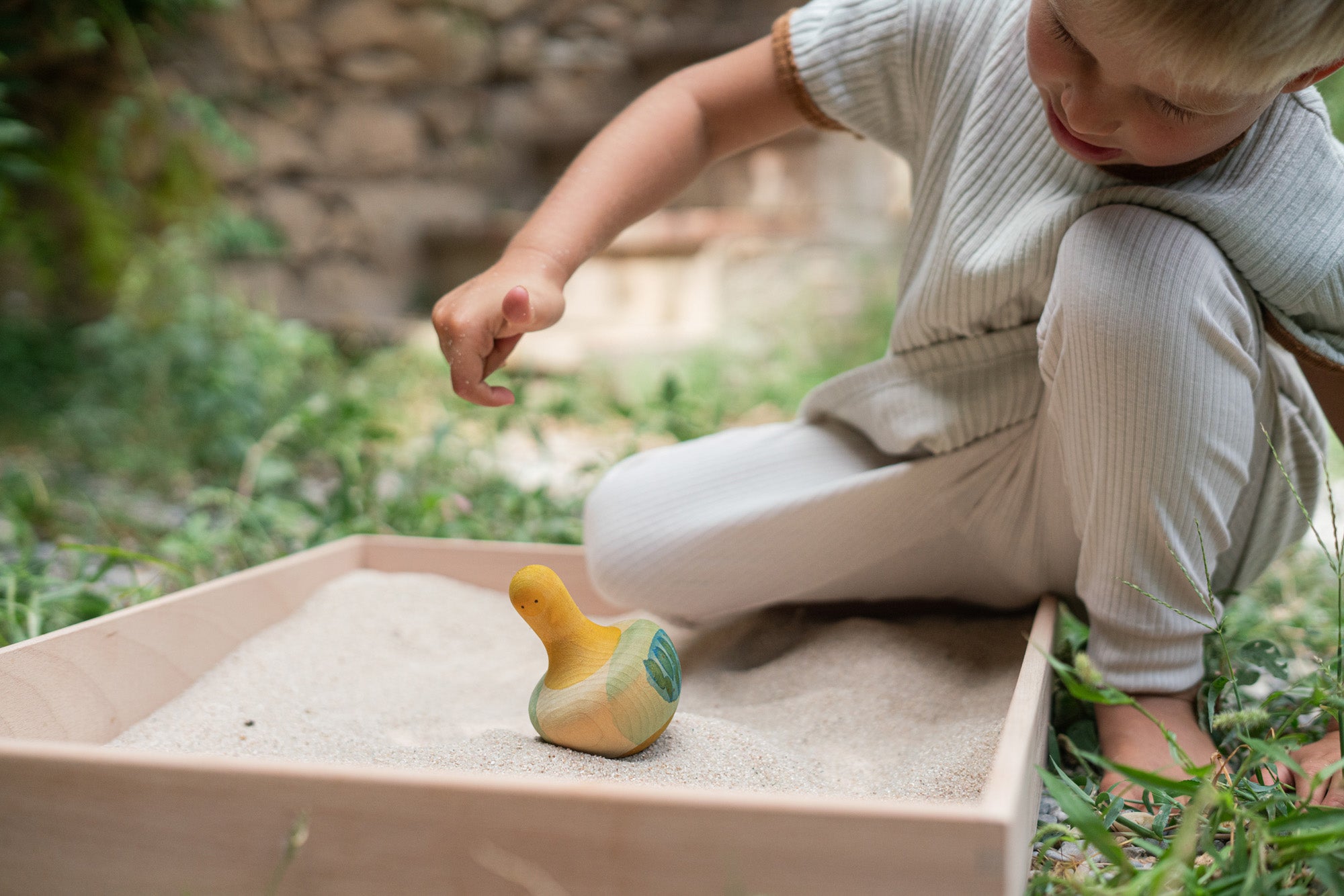 A child in the garden playing with the Grapat wooden yellow flowing bird in a Grapat freeplay tray filled with sand. Showing the hand painted face, blue and green feather detailing