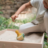 A child in the garden playing with the Grapat wooden yellow flowing bird in a Grapat freeplay tray filled with sand. Showing the hand painted face, blue and green feather detailing