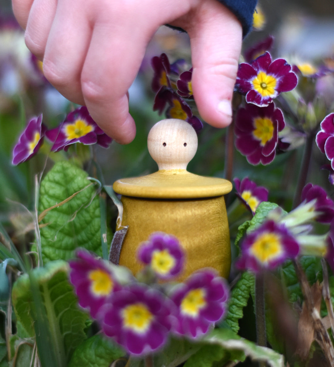 A child reaching for the lid of the Grapat yellow Little Things that has been placed amongst flowers outdoors. A wooden top with eyes drawn on and a yellow coloured pot to match. Grapat have a wide range of wooden toys and loose parts available at Babipur. 