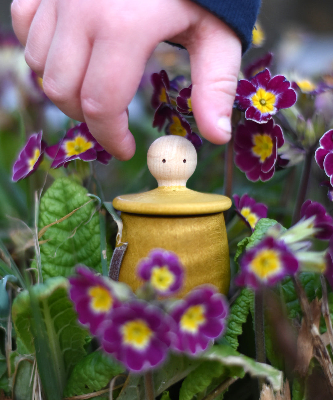 A child reaching for the lid of the Grapat yellow Little Things that has been placed amongst flowers outdoors. A wooden top with eyes drawn on and a yellow coloured pot to match. Grapat have a wide range of wooden toys and loose parts available at Babipur. 