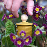 A child reaching for the lid of the Grapat yellow Little Things that has been placed amongst flowers outdoors. A wooden top with eyes drawn on and a yellow coloured pot to match. Grapat have a wide range of wooden toys and loose parts available at Babipur. 